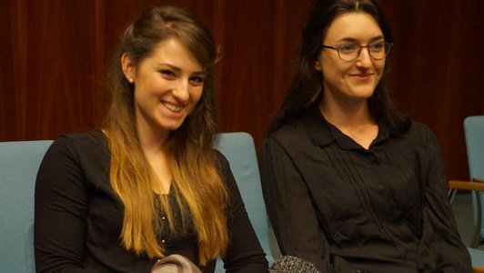 Two women are sitting on blue chairs, smiling.