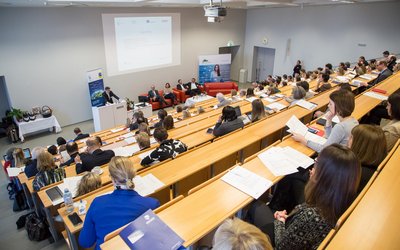 Es wir ein Vortrag in einem Vorlesungssaal gehalten. Es steht ein Man vor einem Podium und redet währenddessen sitzen vier Personen schräg hinter ihm auf einer Couch. Viele Leute sind am Aufpassen und Mitschreiben.