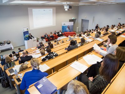 Es wir ein Vortrag in einem Vorlesungssaal gehalten. Es steht ein Man vor einem Podium und redet währenddessen sitzen vier Personen schräg hinter ihm auf einer Couch. Viele Leute sind am Aufpassen und Mitschreiben.