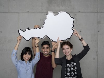 3 internationale Studierende halten eine Österreich Cut-Out in die Luft.