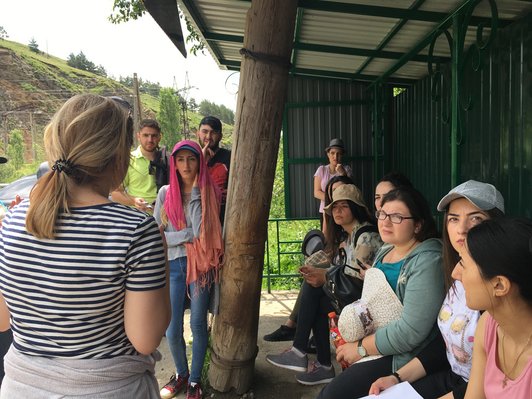 Group of people sitting and standing somewhere outdoors are looking at a woman