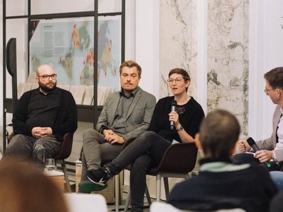 Experten sitzen in einer Halle und diskutieren. Sie spricht in ein Mikrofon, die umstehenden Personen horchen gespannt. Die Wände bestehen aus Marmor.