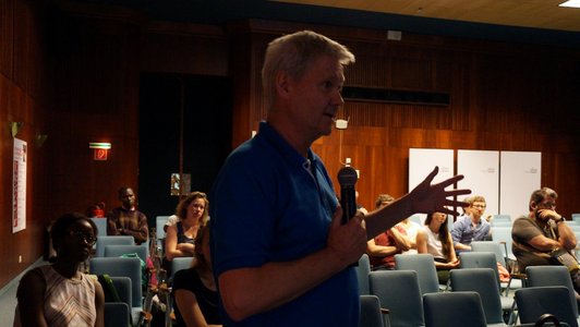 A man is holding a microphone in his hand and speaking in front of a sitting audience. 