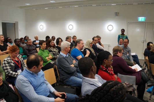 An audience sitting on chairs. One person is standing, one person is speaking into a microphone.