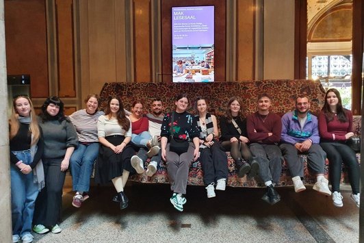 Eine Gruppe von Personen nebeneinander auf einem riesigen Sofa, im Hintergrund ein Bildschirm mit dem Text "MAK Lesesaal"