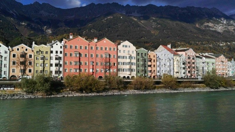 Eine bunte Häuserfront hinter einem Fluss und vor Bergen an einem bewölktem Tag.