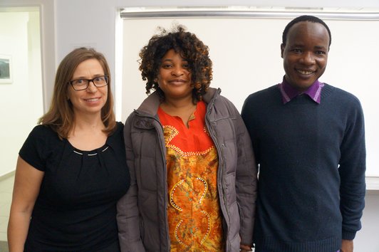 Three people standing in a room with plain white walls, smiling into the camera.