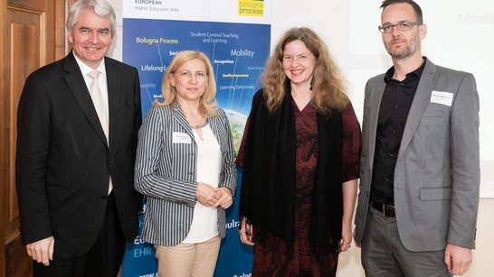 Zwei Männer und zwei Frauen stehen nebeneinander in einem Raum vor einem Rollup und blicken in die Kamera