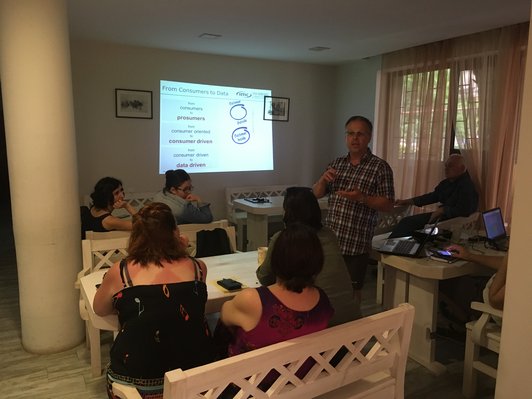 Group of people are looking at a man, who is standing in a darkened room in front of a presentation talking.