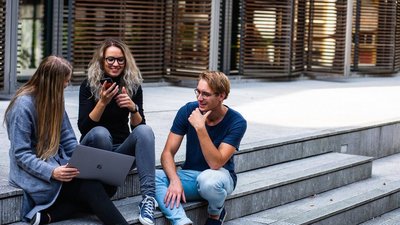 Drei junge Menschen (zwei Frauen und ein Mann) sitzen draußen auf Stufen und unterhalten sich. Eine Frau hält einen Laptop auf dem Schoß, die andere hält ihr Handy in der Hand.