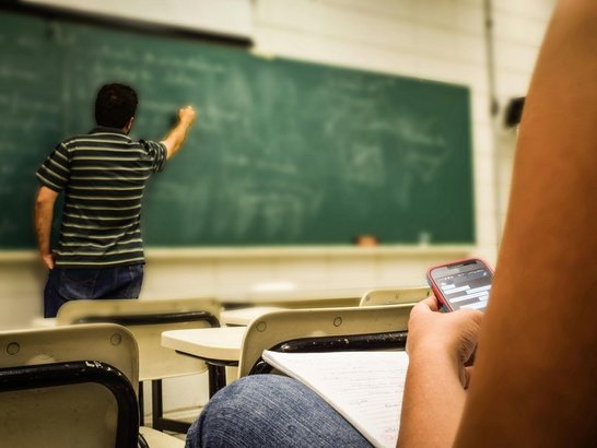 Im Hintergrund schreibt eine Person auf eine grüne Schultafel, im Vordergrund eine sitzende Person von hinten mit Mobiltelefon in der Hand