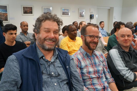 The picture shows an audience sitting on chairs and it is a close-up of three people who are sitting in the front row, smiling.