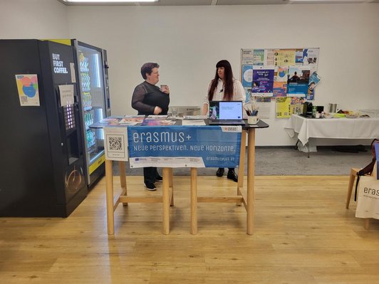 Zwei Menschen stehen an einem Tisch, davor hängt ein türkiser Erasmus+ Banner 