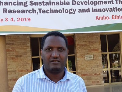 Geda Oncho standing in front of Ambo University, with a big poster in the background