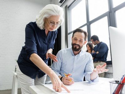 Ältere Dame und junger Herr arbeiten gemeinsam an Dokument vor Bildschirm
