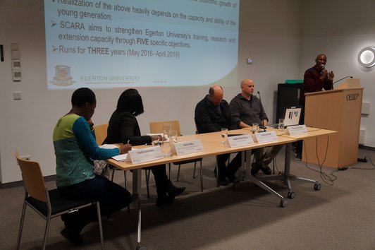 Four people are sitting at a table with name plates, glasses of water, sheets of paper and a laptop on it while one person is giving a speech at a speaker's desk next to them.