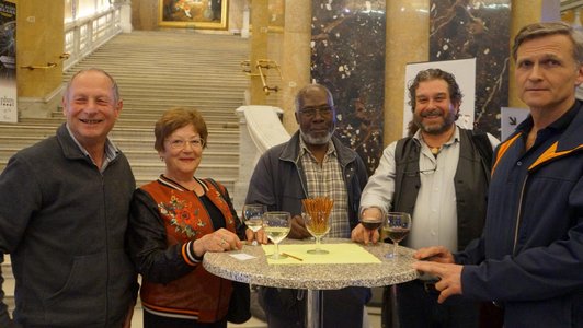 One woman and four men are standing around a table with drinks and pretzel sticks on it. There are marble walls and a beautiful staircase in the background. 