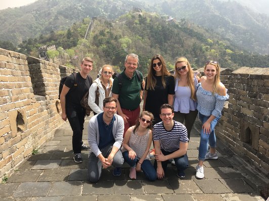 Group of people standing on a large wall and looking at the camera, in the background a forested hill on which the wall continues