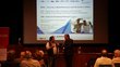 Two people are standing in front of an audience in a lecture hall. One of them is interviewing the other. A large screen in the background shows a presentation. Next to the screen is a red-and-white-coloured roll-up.