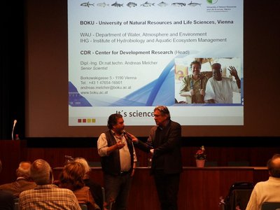 Two people are standing in front of an audience in a lecture hall. One of them is interviewing the other. A large screen in the background shows a presentation. Next to the screen is a red-and-white-coloured roll-up.