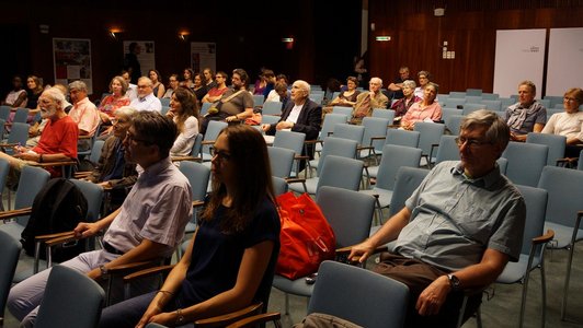 An audience is sitting on blue chairs in a room with wooden walls.