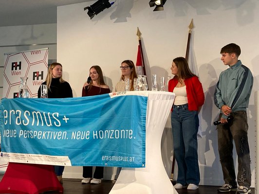 Fünf Personen stehen auf einer Bühne und halten einen türkisen Erasmus+ Banner hoch