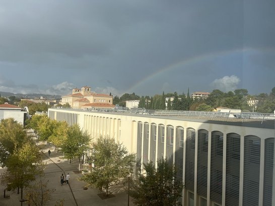 Dunkler Himmel mit Regenbogen, davor ein halb-schattig-halb-sonniges Gebäude