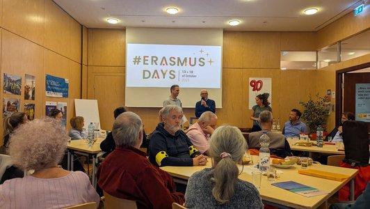 Gruppe an Menschen sitzt an Tischen, auf der Bühne sprechen zwei Männer