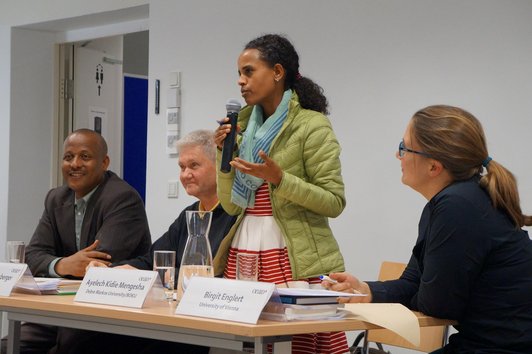 Three people are sitting at a table with name plates, glasses of water, books and sheets of paper on it. One person is standing at the table and speaking into a microphone. 