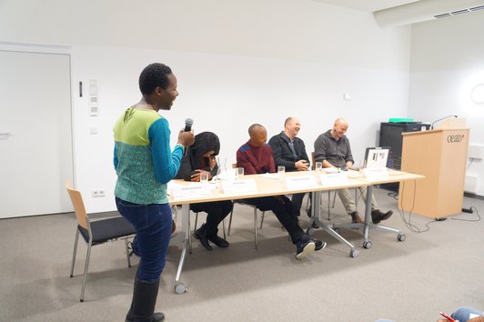 Four people are sitting at a desk with name plates, glasses of water and sheets of paper on it, one person is standing and speaking into a microphone. All of them are laughing cheerily. 