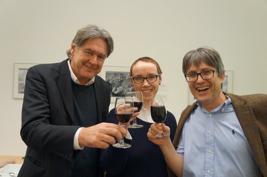 Three people standing together with glasses of wine, smiling into the camera.