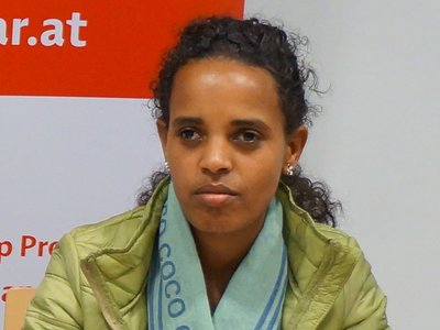 A woman is sitting at a desk, she is having her hands on some sheets of paper and there is a glass of water standing in front of her.