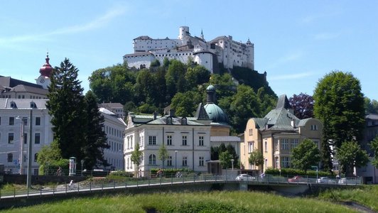 Eine weiße Festung auf einem bewaldeten Berg über einigen historischen Häusern. Im Vordergrund ist ein Fluss.