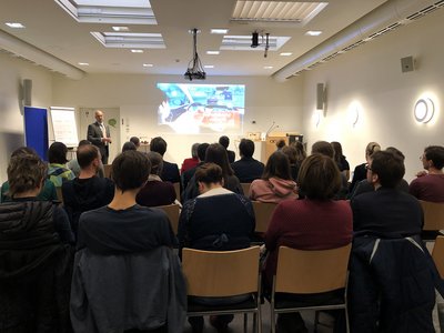 Martin Bauer hält einen Vortrag vor Leinwand, man sieht das Publikum von hinten