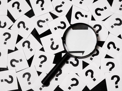 Zettel mit Fragezeichen und eine Lupe, die darauf gerichtet ist