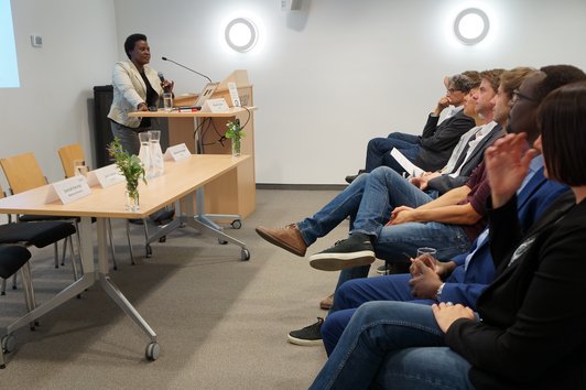 Group of people looking at a woman, who is standing behind a desk presenting the project