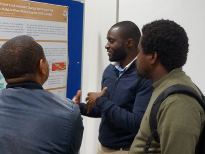 Three men standing in front of a poster of the poster presentation discussing its content