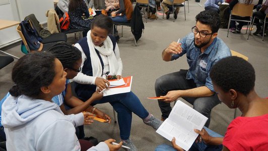 Five people sitting in chairs in a circle and discussing, with more people in the background