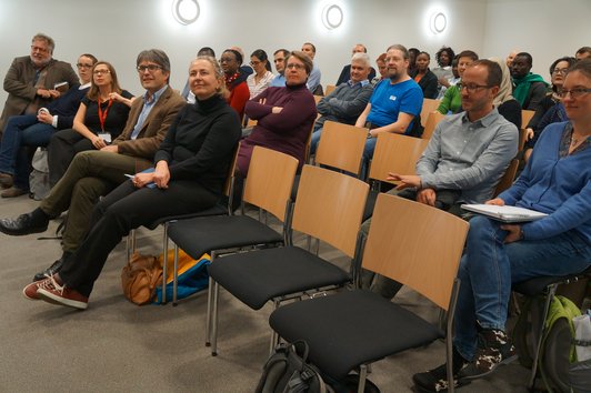 An audience sitting on chairs in a room with white walls and a grey carpeted floor.