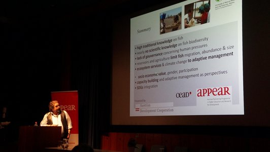 A man is standing at a speaker's desk and looking up at a large screen. The logos of the appear programme, of the BOKU, the OeAD and the Austrian Development Cooperation are shown on the presentation that is projected onto the wall.