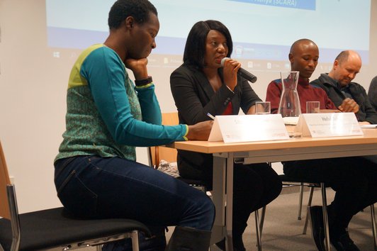 People sitting at a desk with name plates and glasses of water on it, in front of a large screen. One of the women is holding a microphone, apparently speaking to the audience. 