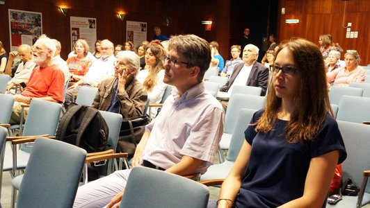 An audience is sitting on blue chairs in a room with wooden walls. There are approximately 30-40 people on the picture.