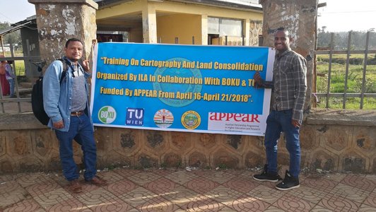 Two man standing next to a big event poster attached to a fence in front of a building