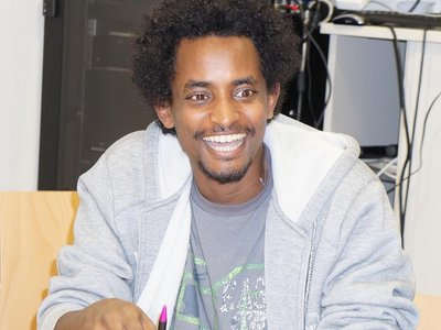 A man sitting at a desk, a pen in his hands and sheets of paper in front of him. He is laughing cheerily.