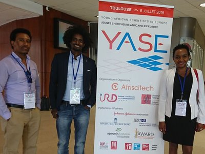 One woman and two men with badges on lanyards around their necks, standing next to a roll-up announcing the "YASE, Young African Scientists in Europe" on 6 July 2018 