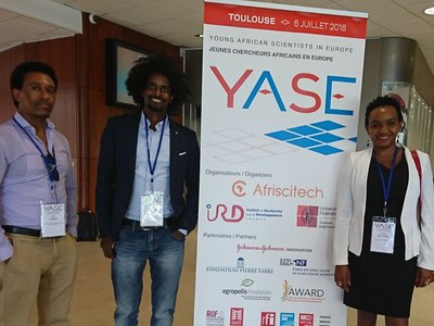 One woman and two men with badges on lanyards around their necks, standing next to a roll-up announcing the "YASE, Young African Scientists in Europe" on 6 July 2018 