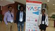 One woman and two men with badges on lanyards around their necks, standing next to a roll-up announcing the "YASE, Young African Scientists in Europe" on 6 July 2018 