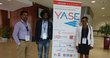 One woman and two men with badges on lanyards around their necks, standing next to a roll-up announcing the "YASE, Young African Scientists in Europe" on 6 July 2018 