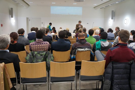 An audience sitting on chairs and following the speech of a person who is standing at the front, speaking into a microphone. 