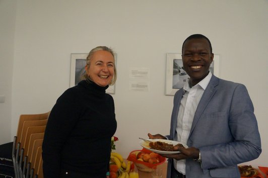 Two people standing and smiling into the camera. One of them is holding a plate with some dish on it.